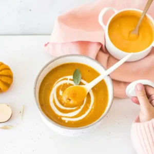 Creamy vegan pumpkin and red lentil soup with a coconut cream swirl, served in a white bowl with a pink napkin and mini pumpkin.