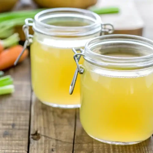 Homemade chicken broth stored in clear glass jars on a wooden table with fresh carrots and green onions in the background.