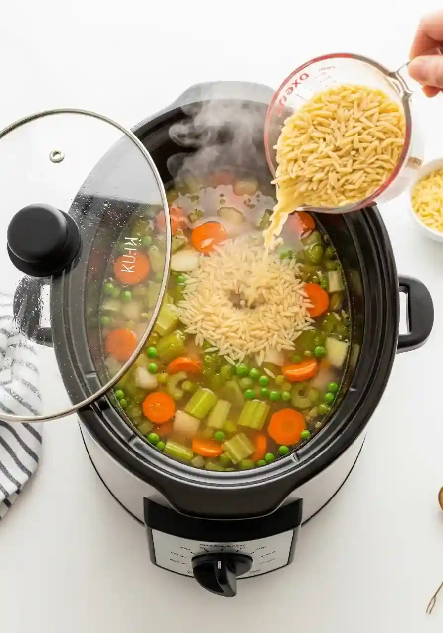 Uncooked orzo being poured into hot broth inside a black oval crockpot.
