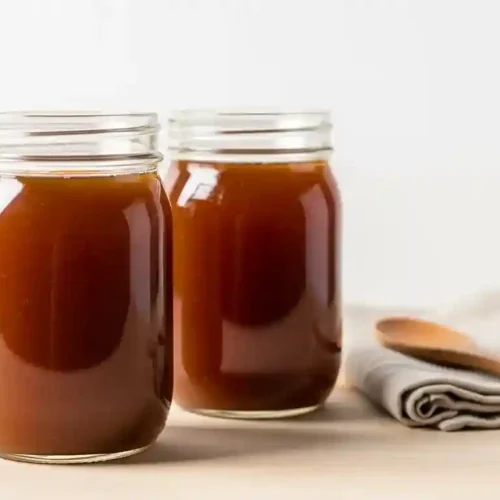 Two glass jars filled with rich dark brown homemade beef broth on a light wooden surface with a white background.