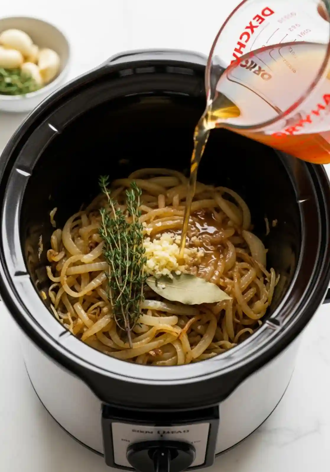 crockpot-french-onion-soup-broth Beef broth being poured into a slow cooker with caramelized onions, garlic, fresh thyme, and bay leaf for crockpot French onion soup