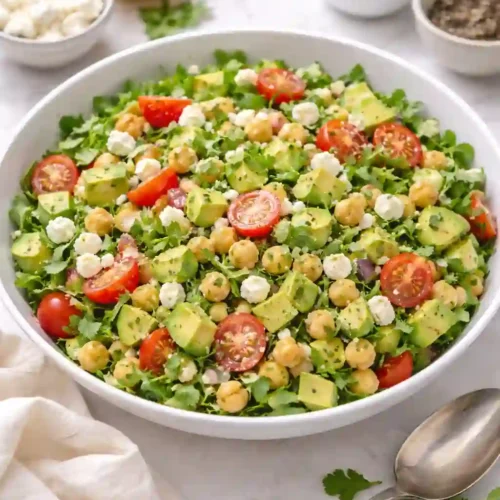Overhead view of chickpea feta avocado salad with tomatoes, arugula, and lemon dressing in a white bowl.