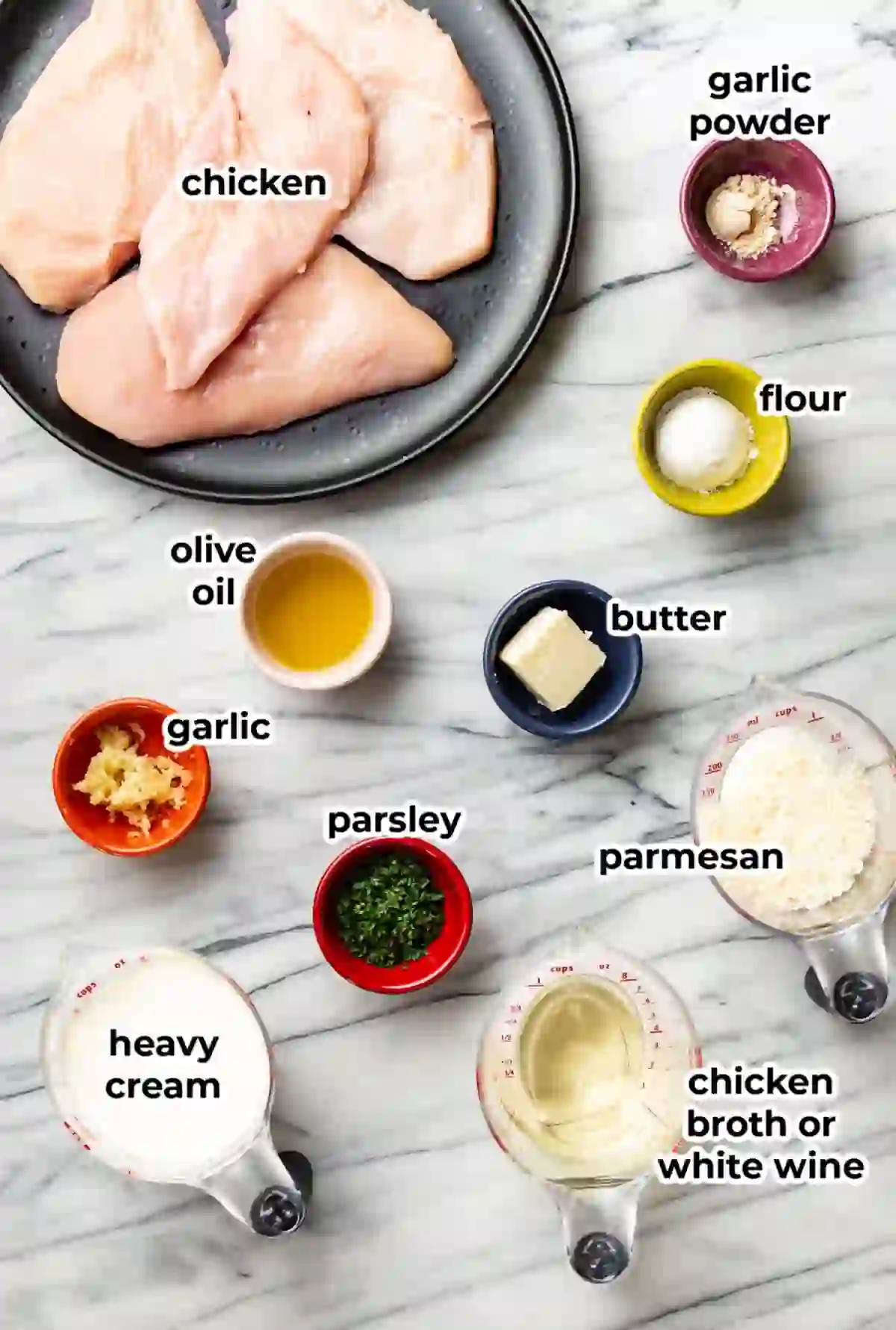 Ingredients for garlic parmesan chicken arranged on a light marble background, including chicken breasts, heavy cream, parmesan, garlic, butter, olive oil, flour, parsley, and chicken broth.