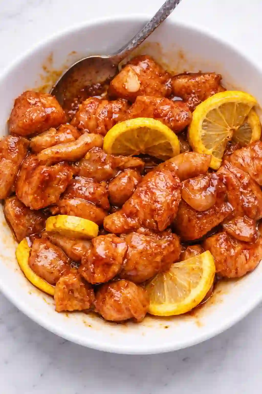 Seasoned chicken pieces tossed with spices and lemon slices in a white bowl on a white table background