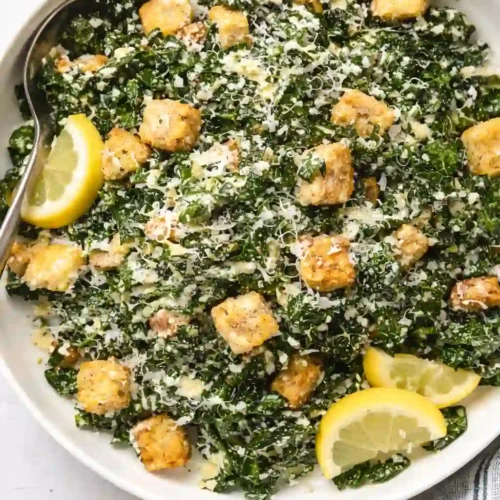 Close-up overhead view of kale caesar salad with chicken, croutons, lemon wedges, and a single spoon in a white bowl