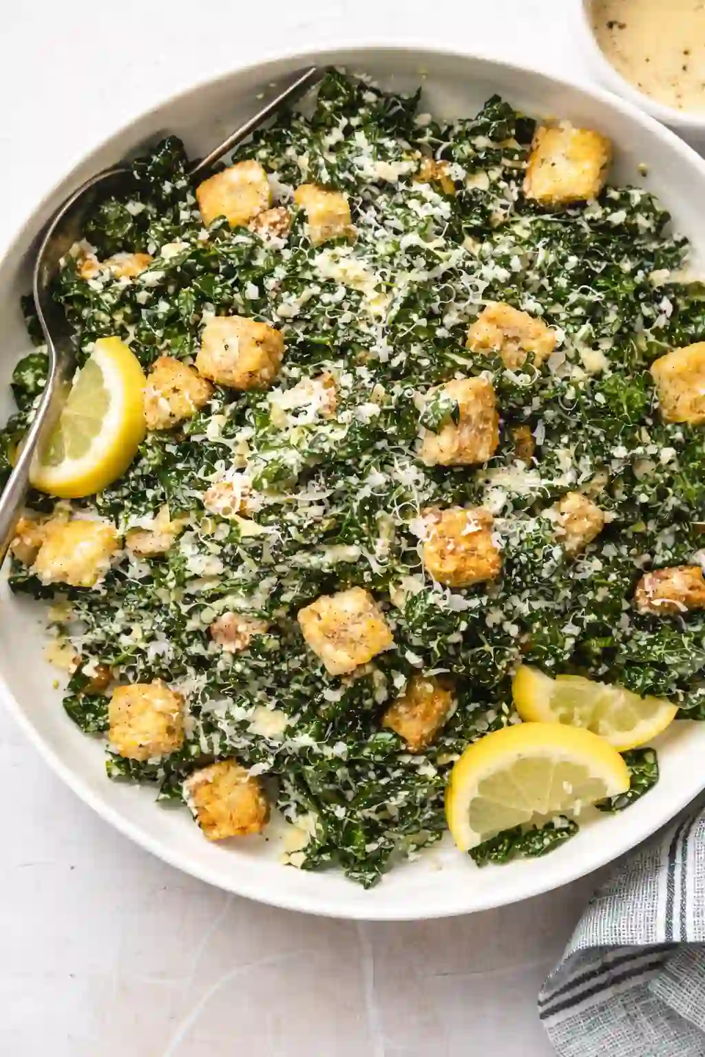 Close-up overhead view of kale caesar salad with chicken, croutons, lemon wedges, and a single spoon in a white bowl