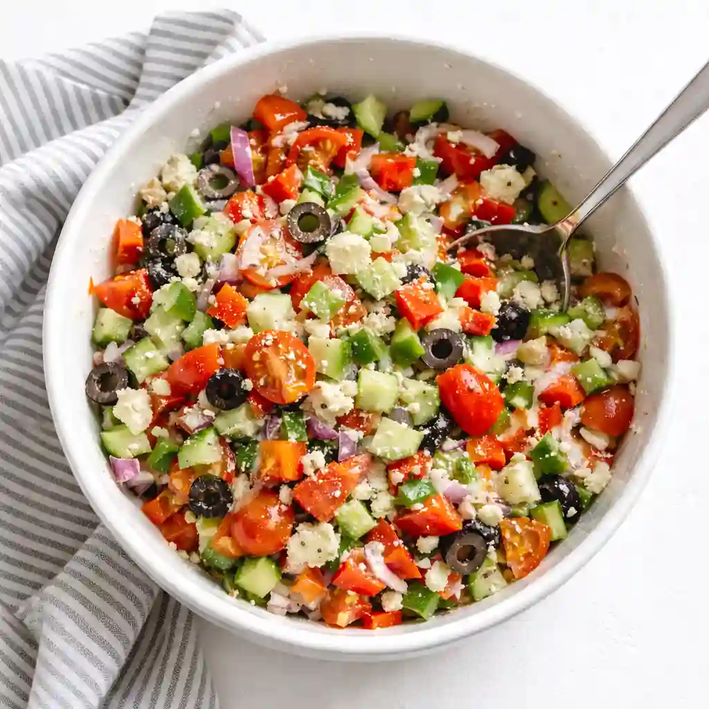 Mixing fresh vegetables, olives, feta, and dressing in a white bowl on a white table background