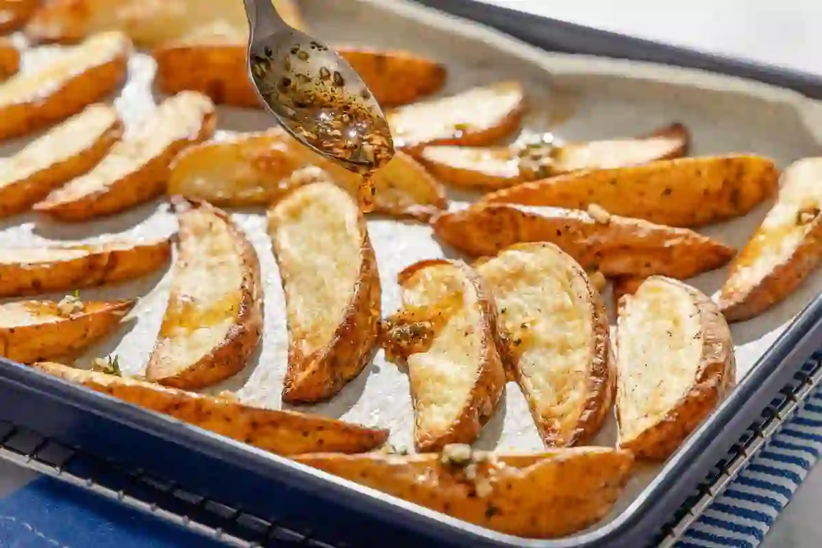 Garlic herb olive oil being spooned over roasted potato wedges on a sheet pan.