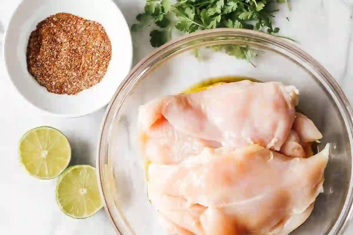 grilled-chicken-breast-ingredients_compressed (1) Raw boneless skinless chicken breasts in a glass bowl with olive oil, lime halves, cilantro, and seasoning on white background