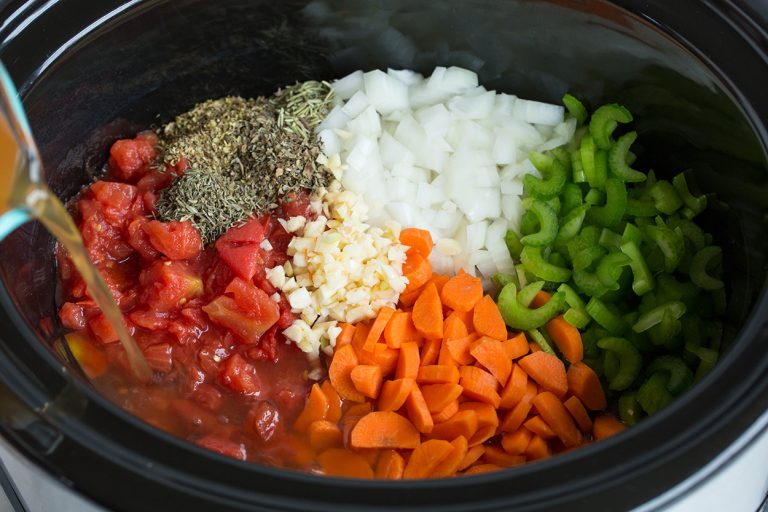 Slow cooker filled with diced tomatoes, carrots, celery, onion, garlic, herbs, and vegetable broth for minestrone soup.