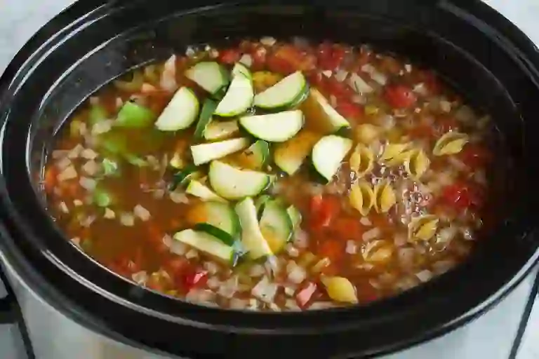 Adding pasta, zucchini, and green beans to minestrone soup in a slow cooker.