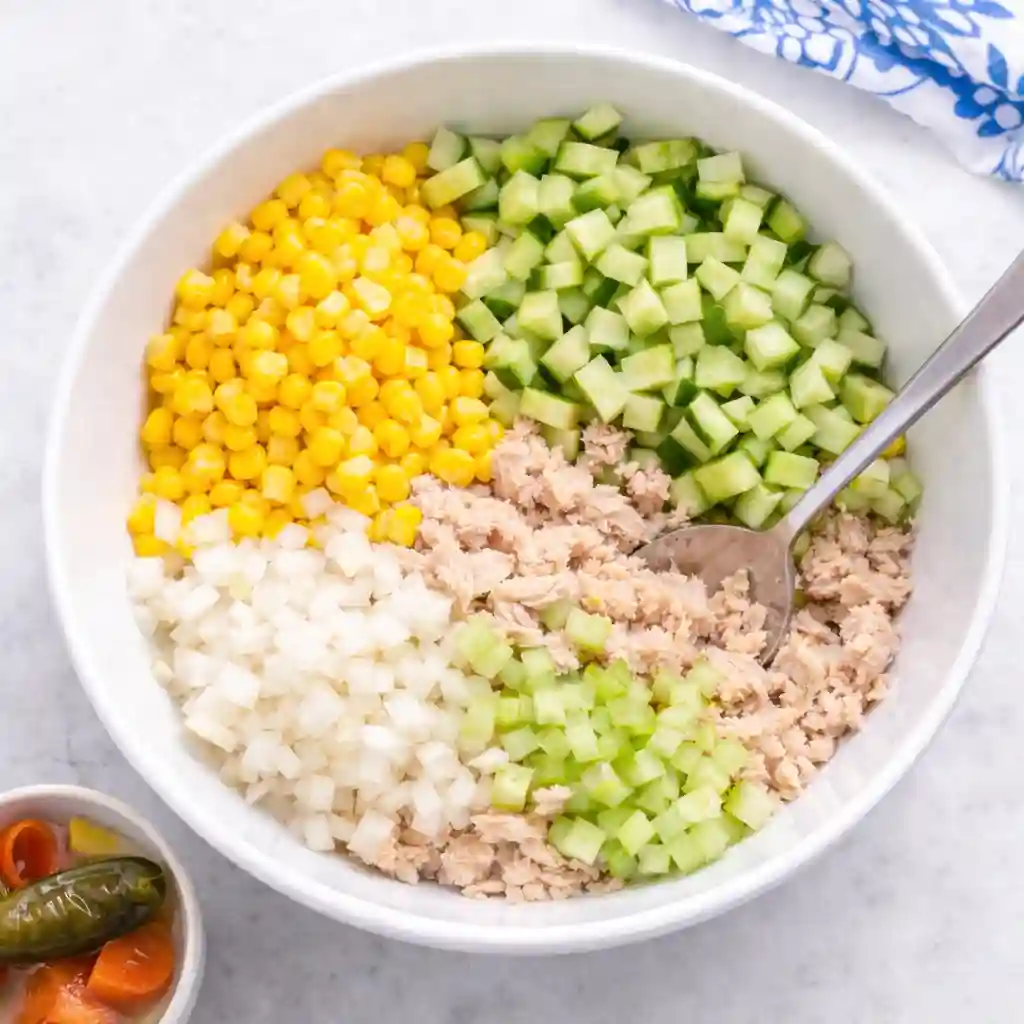 Adding corn, cucumber, onion, and celery to tuna while preparing mexican tuna salad recipe with mayonnaise.