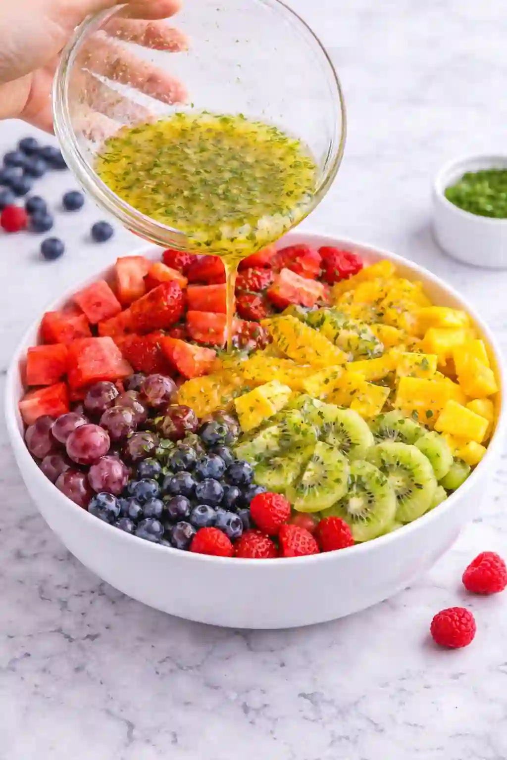 step-3-pour-dressing-fruit-salad-vertical._compressed Pouring honey lime dressing over fresh fruit salad in a white bow