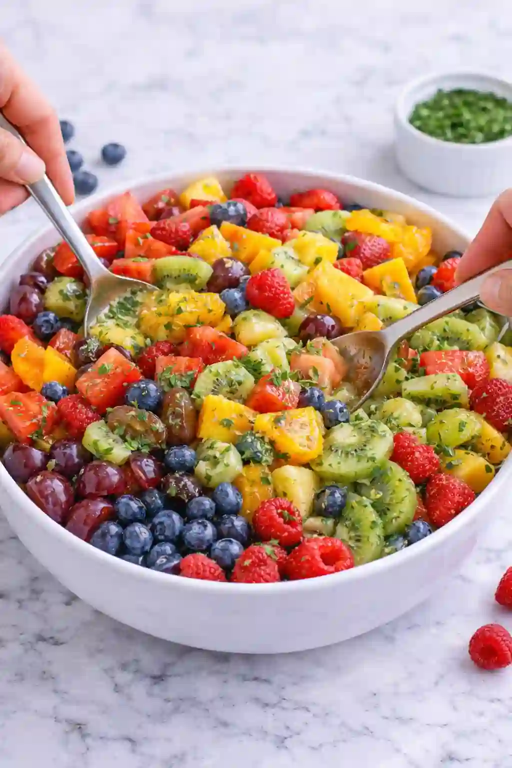 step-4-mix-fruit-salad-vertical._compressed (1) Mixing fresh fruit salad in a white bowl with spoon and fork