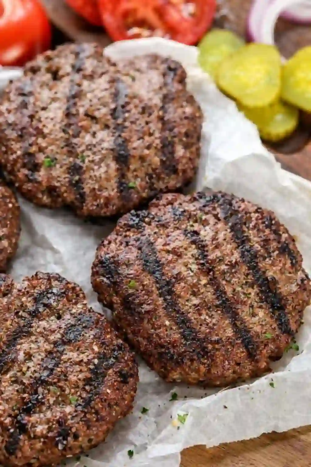  grilled hamburger patties with grill marks resting on parchment paper with pickles and tomatoes
