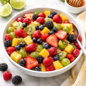 Fresh fruit salad in a white bowl with strawberries, kiwi, pineapple, grapes, blueberries, and raspberries coated in a light honey lime dressing on a bright clean background