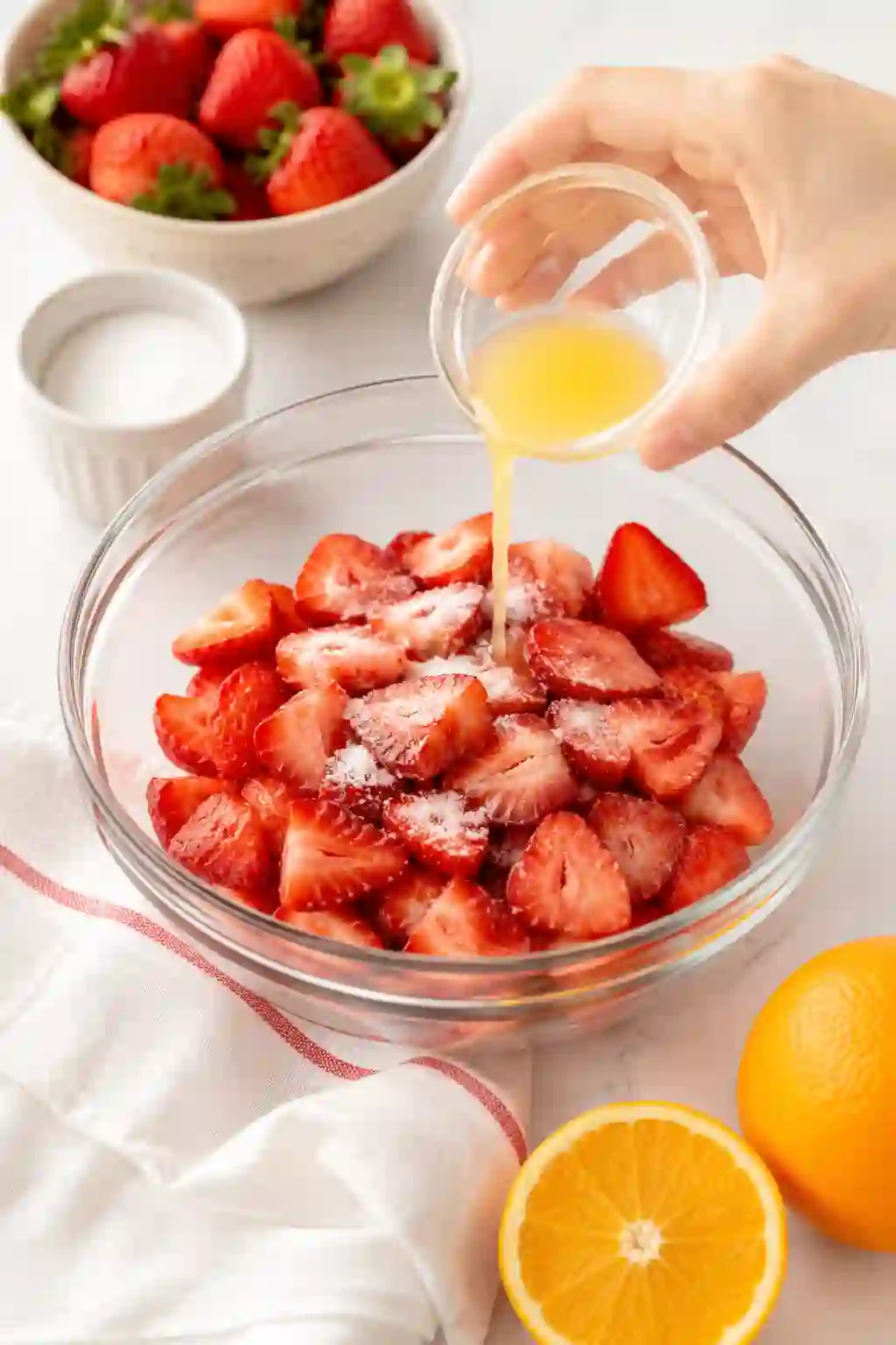 Fresh sliced strawberries in a glass bowl with sugar and orange juice being poured over them on a bright kitchen surface